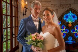 Portrait d'un couple de mariés souriants, la mariée en robe blanche tenant un bouquet de fleurs, posant devant une grande fenêtre en bois avec vitraux colorés en arrière-plan.