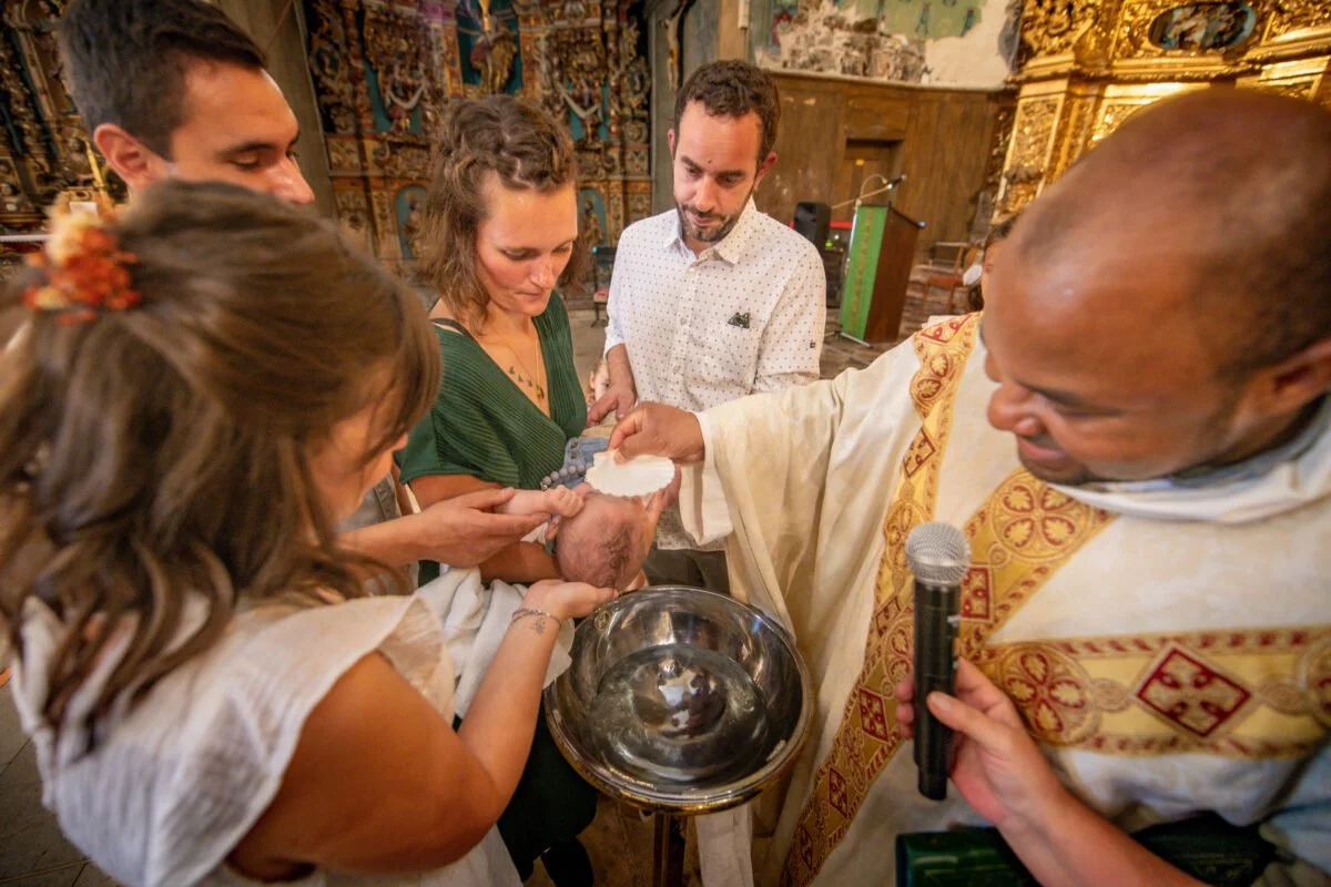 Scène de baptême en église, avec le prêtre versant l’eau sur la tête du bébé, entouré par les parents et parrains.