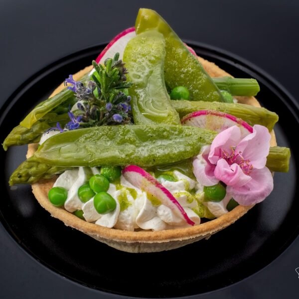 Tartelette salée aux légumes verts et fleurs comestibles, dressée sur assiette noire, photographiée en gros plan.