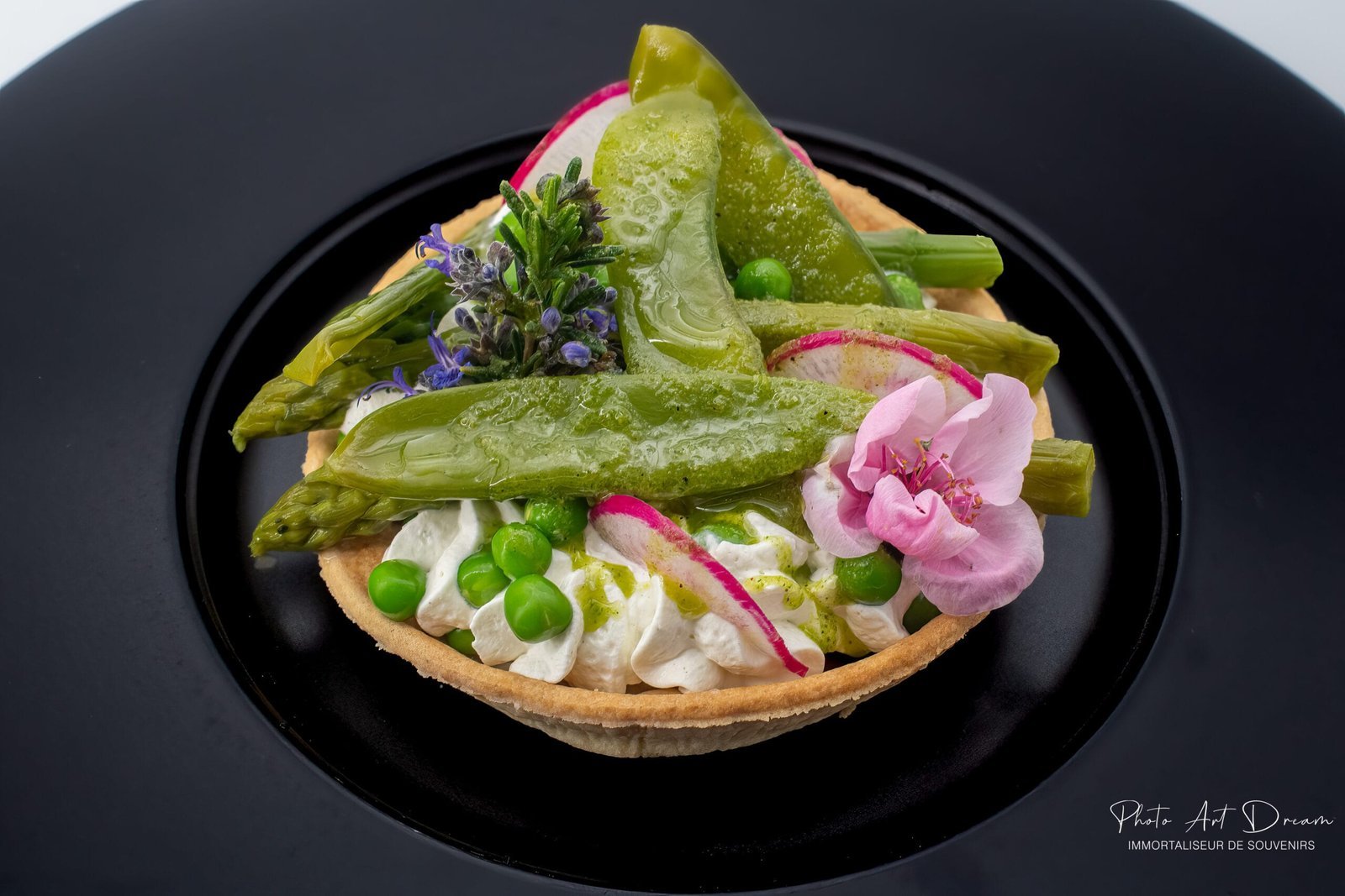 Tartelette salée aux légumes verts et fleurs comestibles, dressée sur assiette noire, photographiée en gros plan.