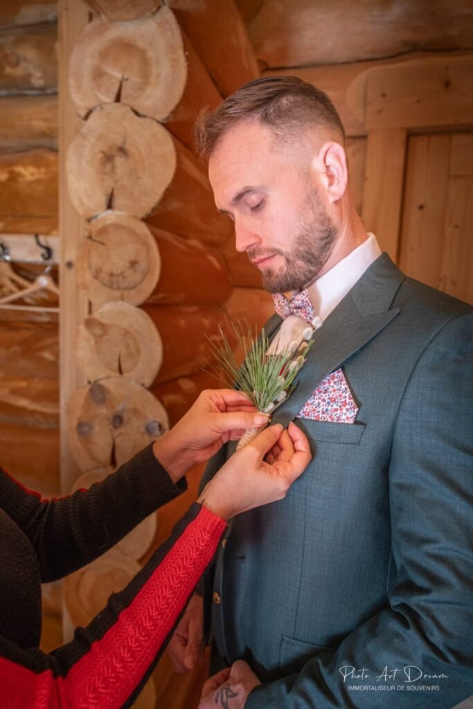 Marié en costume vert sapin recevant sa boutonnière florale dans un chalet en bois à Mosset.