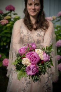 Mariée tenant un bouquet de pivoines roses et de roses crème devant un massif d’hortensias fleuris.