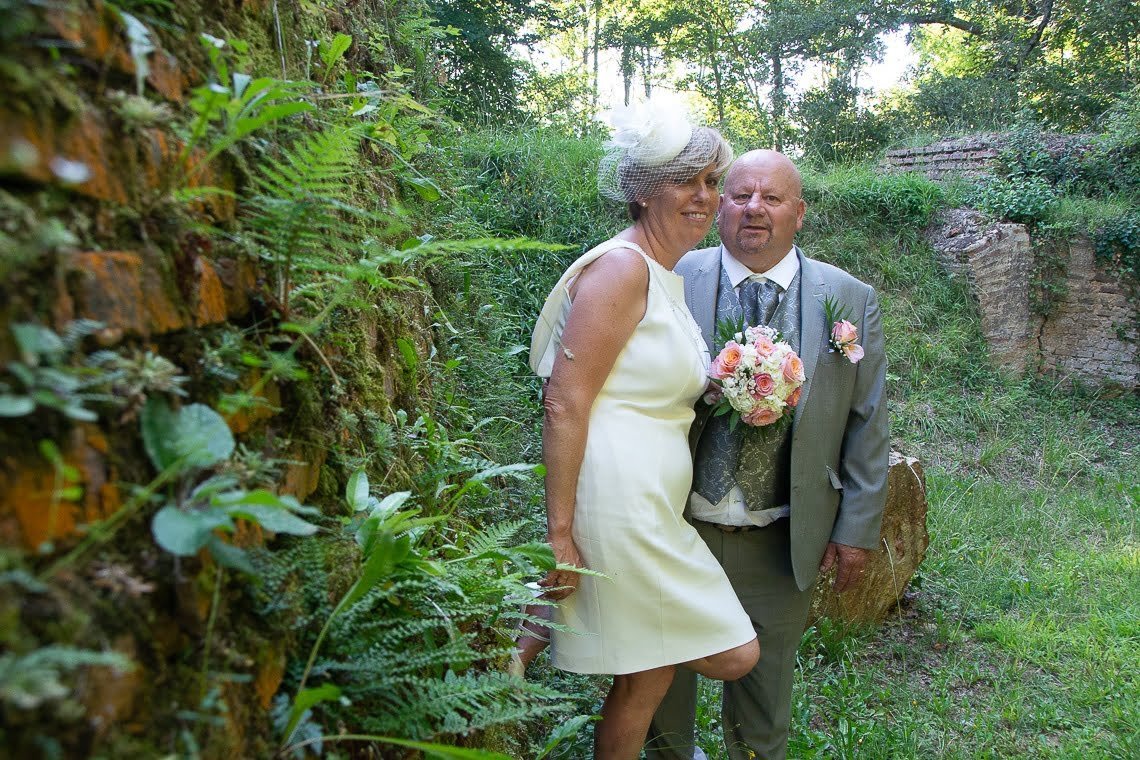 Couple de mariés posant dans un décor naturel avec mur en pierre recouvert de mousse, la mariée en robe courte ivoire et chapeau vintage.