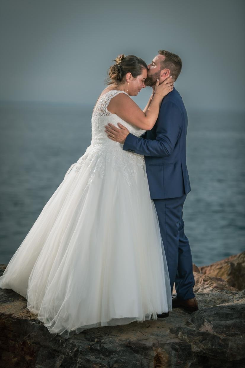 Mariés s’enlaçant tendrement sur les rochers, au bord de la mer, le marié embrassant le front de la mariée en robe blanche.