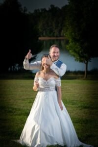Mariés souriants dans un parc, le marié cache les yeux de la mariée en faisant un signe de pouce levé.