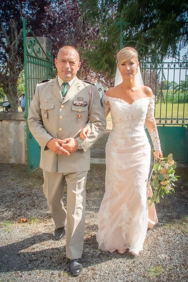 Mariée en robe en dentelle marchant au bras d’un militaire en uniforme beige, à l’entrée d’un domaine verdoyant.