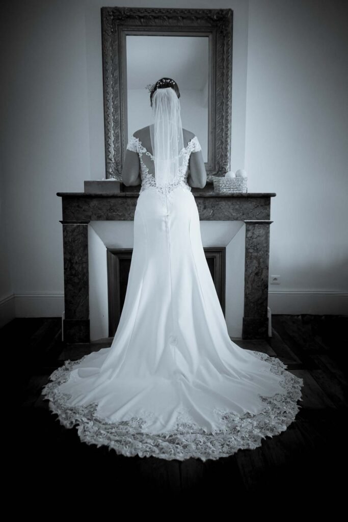Photographe mariage à Perpignan - Mariée de dos en robe longue avec traîne en dentelle, devant une cheminée décorative à Thuir, capturée en noir et blanc