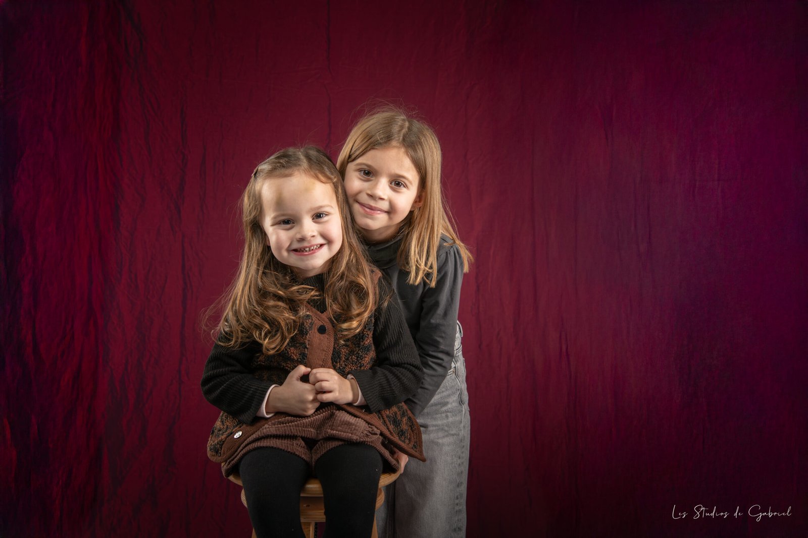 Portrait de deux sœurs souriantes en studio, assises ensemble sur fond rouge, dans une ambiance douce et naturelle
